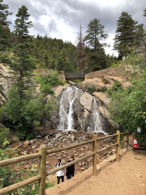 A serene waterfall cascades over rocks, surrounded by lush greenery and tall trees, with a path and wooden fence in view.