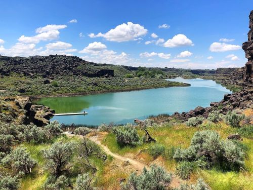 A serene landscape featuring a calm lake surrounded by rocky hills and lush greenery under a blue sky with fluffy clouds.