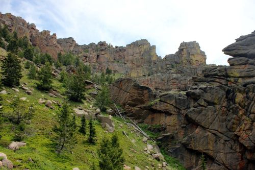 Rocky cliffs rise above a lush green hillside, dotted with trees and boulders under a cloudy sky.