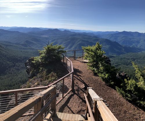 A scenic overlook with wooden railings, surrounded by lush green mountains under a clear blue sky.