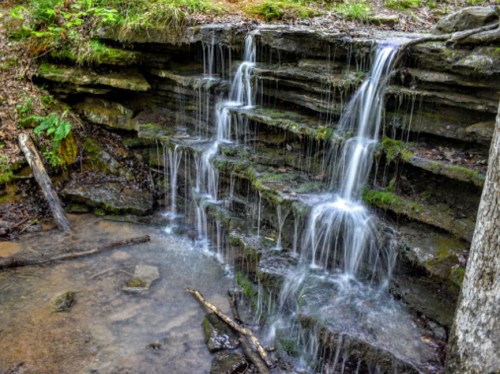 A serene waterfall cascading over layered rocks into a calm pool, surrounded by lush greenery.