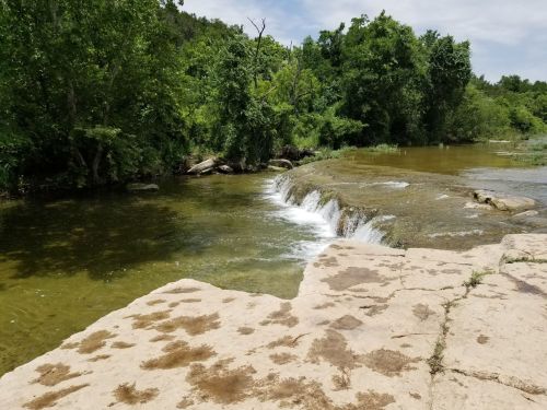 A serene river scene with a small waterfall, surrounded by lush green trees and a rocky shoreline.