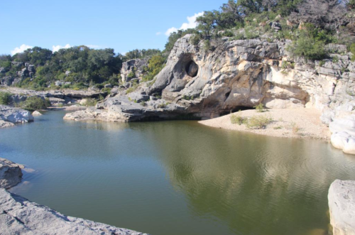A serene river scene with rocky cliffs and lush greenery under a clear blue sky. Reflections shimmer on the water's surface.