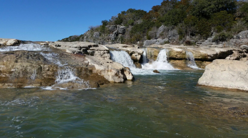 A serene landscape featuring cascading waterfalls over rocky terrain, surrounded by lush greenery and a clear blue sky.