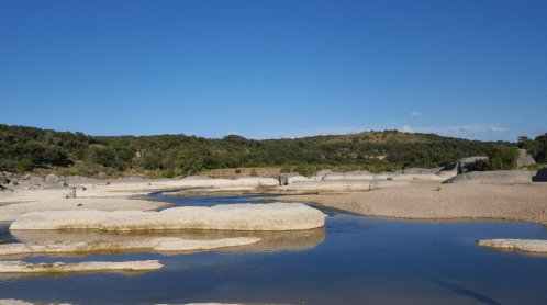 A serene landscape featuring smooth rocks and a calm river under a clear blue sky with distant hills.