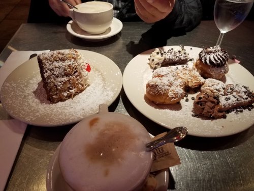 A table with a cappuccino, a slice of cake, and an assortment of pastries dusted with powdered sugar.