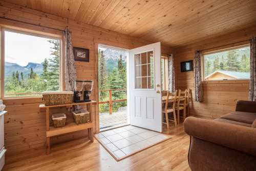 Cozy wooden cabin interior with large windows, a door to a deck, and a dining area surrounded by nature.
