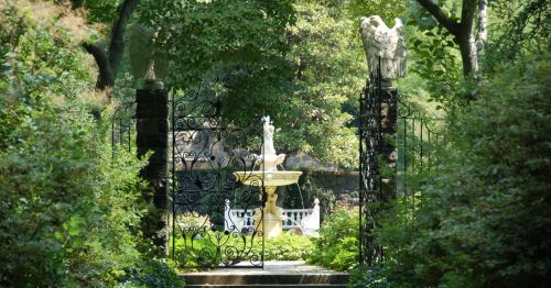 A lush garden entrance with ornate iron gates, flanked by stone sculptures, leading to a fountain surrounded by greenery.