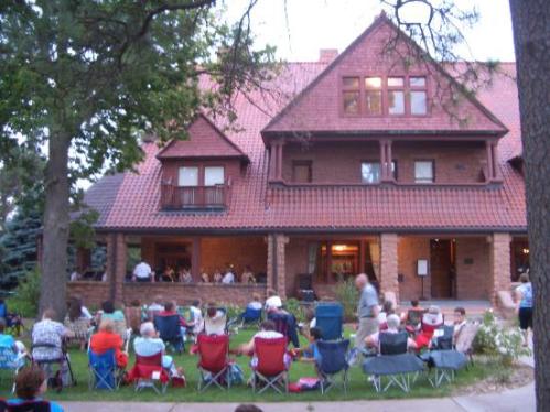 A large gathering of people sitting in lawn chairs in front of a historic brick building with a red roof.