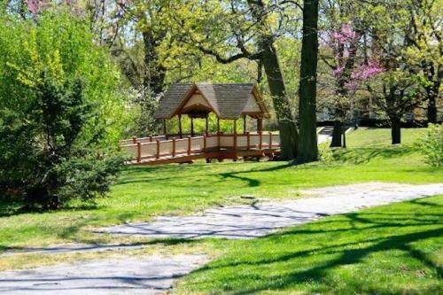 A wooden gazebo on a grassy area, surrounded by trees and blooming flowers in a sunny park.