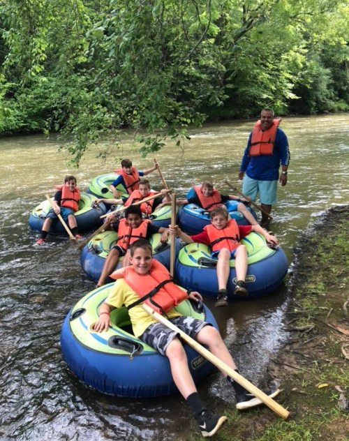 A group of children in colorful inner tubes on a river, wearing life jackets and smiling, with a guide nearby.