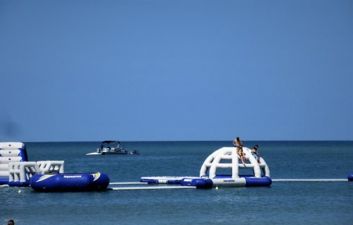 Two children play on a floating water structure in a calm sea, with a boat in the background under a clear blue sky.