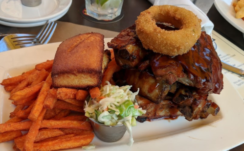 A plate of ribs topped with barbecue sauce, served with sweet potato fries, coleslaw, cornbread, and an onion ring.