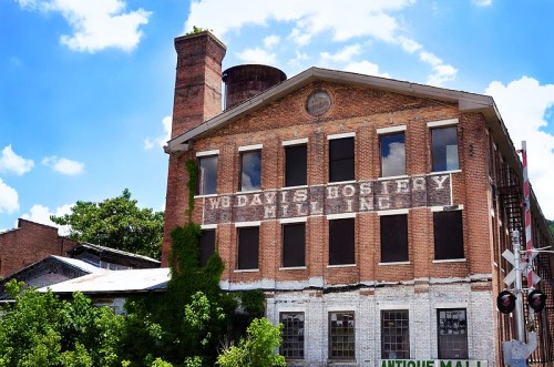 Historic brick building with large windows, featuring faded signage for "W. D. Davis Hosiery Mill" and greenery.