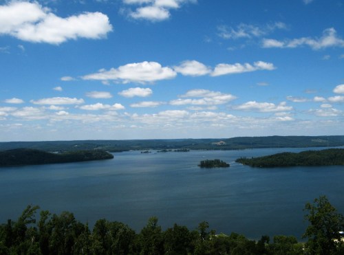 A serene lake surrounded by green hills under a blue sky with fluffy white clouds.