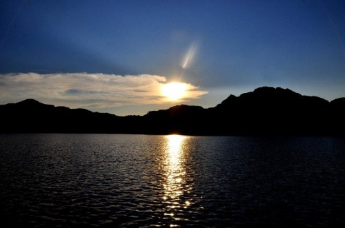 Sunset over a calm lake, with silhouettes of mountains and a reflective water surface under a blue sky.