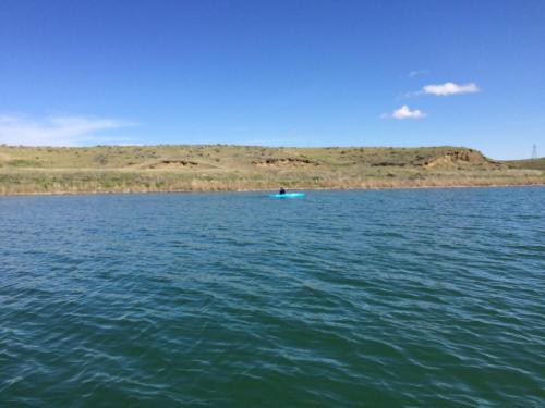 A person kayaking on a calm lake with green hills and a clear blue sky in the background.