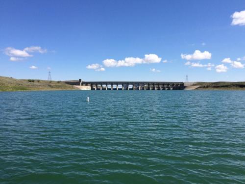 A view of a dam across a calm lake, with blue skies and scattered clouds in the background.