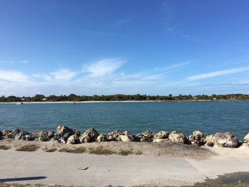 A calm river scene with rocky shoreline, blue water, and a clear sky with scattered clouds.