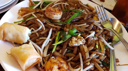 A plate of stir-fried noodles with shrimp, green onions, bean sprouts, and spring rolls on the side.