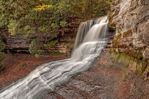 A serene waterfall cascading over rocky terrain, surrounded by lush greenery and autumn foliage.