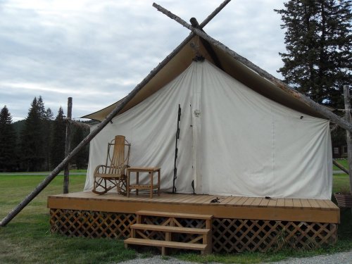 A canvas tent on a wooden deck, with a rocking chair and a chair outside, set in a grassy area surrounded by trees.