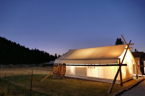 A lit canvas tent stands in a grassy area at dusk, surrounded by trees and a starry sky.
