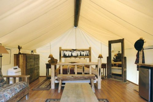 Cozy glamping tent interior with a wooden bed, dresser, lamps, and rustic decor on wooden floors.