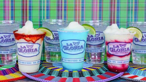 Colorful frozen drinks in cups with lime slices, displayed on a vibrant woven mat, with a checkered background.