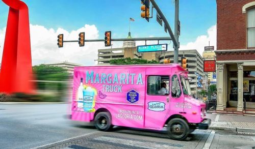 A pink margarita truck drives through an intersection, with a cityscape and traffic lights in the background.