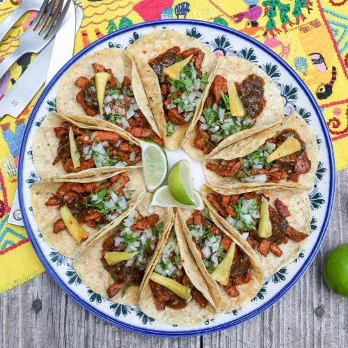 A colorful plate of tacos topped with meat, onions, and pineapple, surrounded by lime wedges on a vibrant tablecloth.