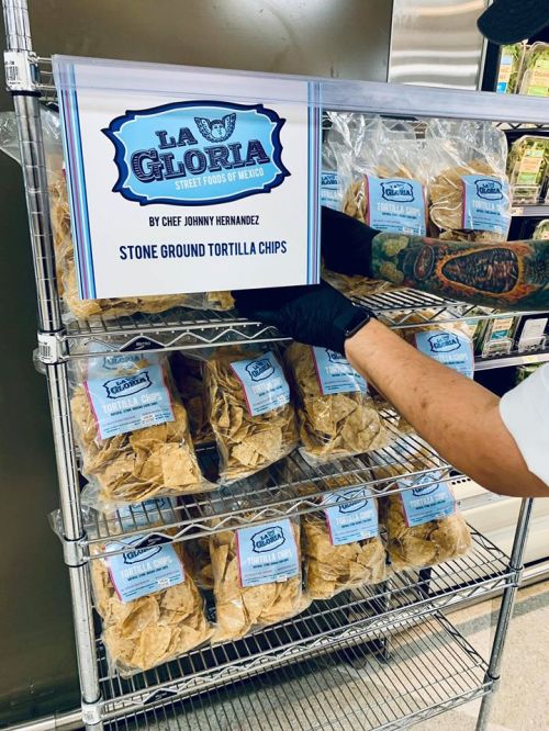 A person in a black glove arranges bags of stone ground tortilla chips on a metal shelf labeled "La Gloria."