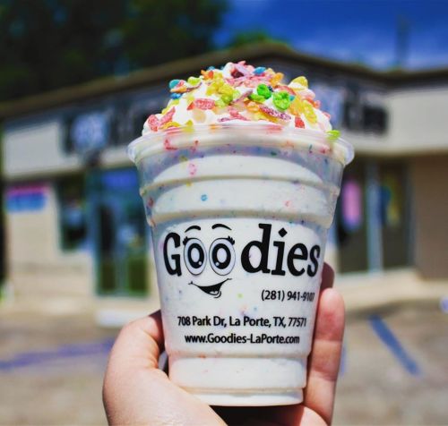 A hand holding a cup of colorful ice cream topped with sprinkles, in front of a shop with a sign that reads "Goodies."