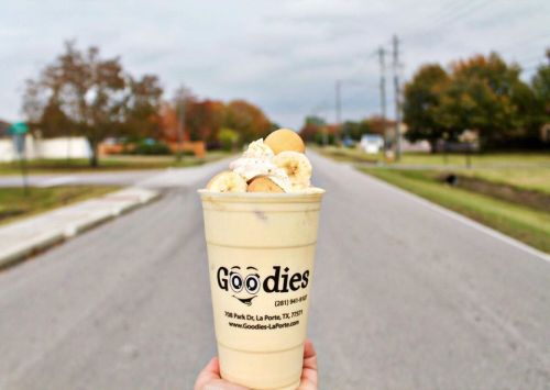 A hand holds a large cup of banana pudding topped with whipped cream and cookies, set against a cloudy street backdrop.