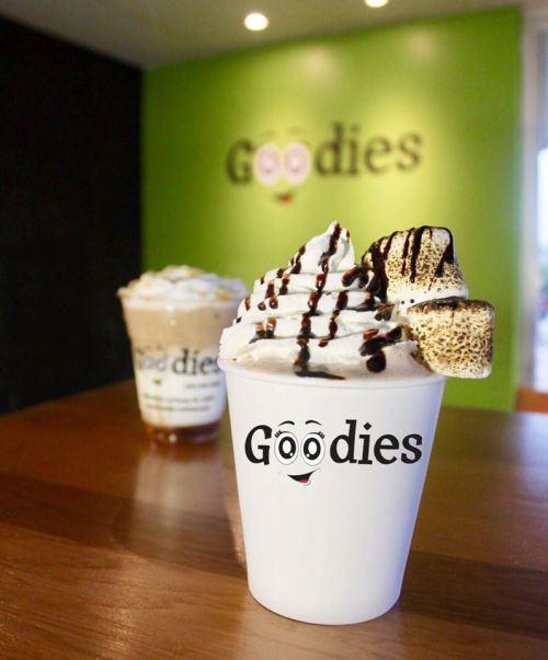 A cup of whipped cream topped with chocolate drizzle and marshmallows, with a blurred drink in the background.