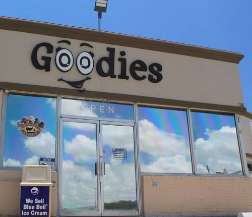 A storefront with the name "Goodies" featuring a cartoonish face, open sign, and colorful reflective windows.