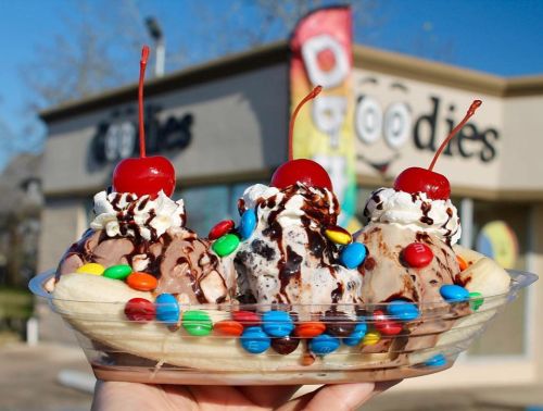 A hand holding a banana split with three scoops of ice cream, topped with whipped cream, chocolate sauce, and cherries.