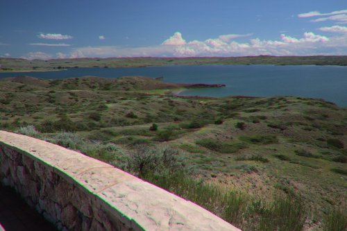 Scenic view of a lake surrounded by green hills under a blue sky with clouds. A stone wall is in the foreground.