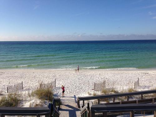 A serene beach scene with white sand, gentle waves, and two people walking along the shore under a clear blue sky.