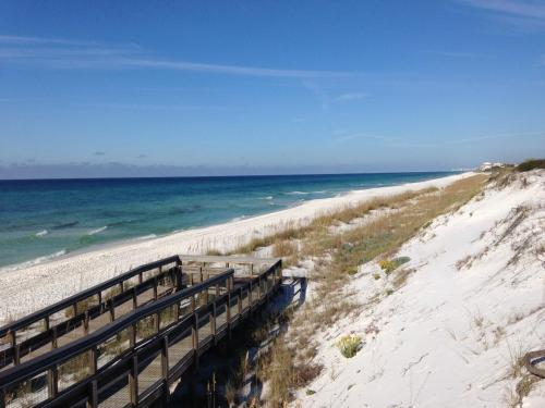 A wooden boardwalk leads to a serene beach with white sand and clear blue water under a bright sky.