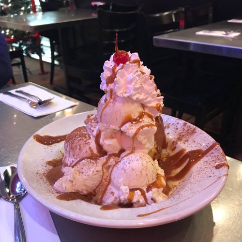 A tall dessert with three scoops of ice cream, whipped cream, caramel drizzle, and a cherry on top, served in a bowl.