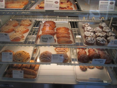 A display case filled with various pastries, including donuts, strudels, and cinnamon rolls, all labeled with prices.