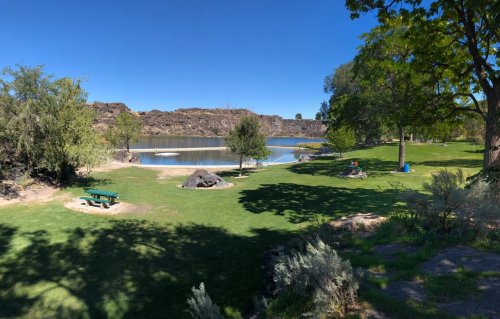 A serene park scene featuring a calm lake, green grass, trees, and picnic areas under a clear blue sky.