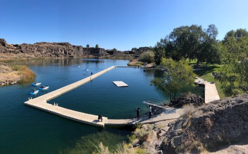 A serene lake with a wooden dock, surrounded by rocky cliffs and greenery, with people kayaking and enjoying the outdoors.