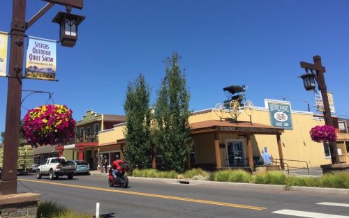 A street view of a quaint town with shops, hanging flower baskets, and a cyclist passing by.