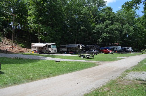 A campsite with several RVs parked among trees, with a gravel road and green grass in the foreground.