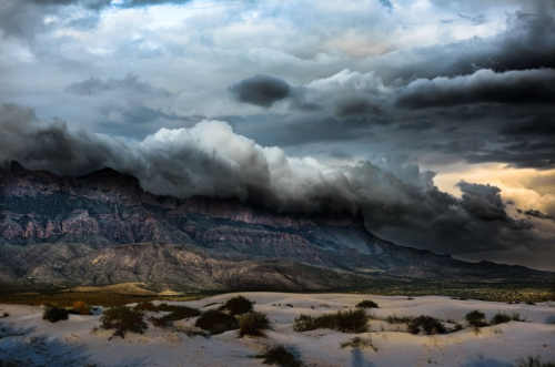 Dramatic landscape featuring dark clouds over rugged mountains and sandy terrain, creating a moody atmosphere.