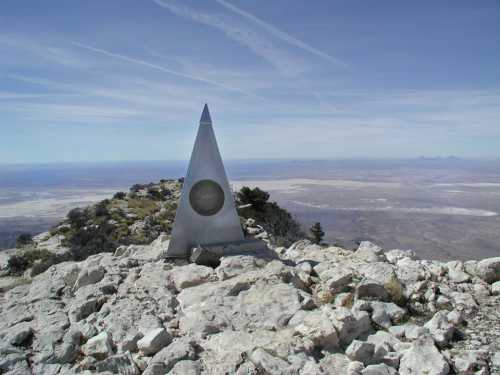 A triangular monument stands on rocky terrain, overlooking a vast, arid landscape under a clear blue sky.