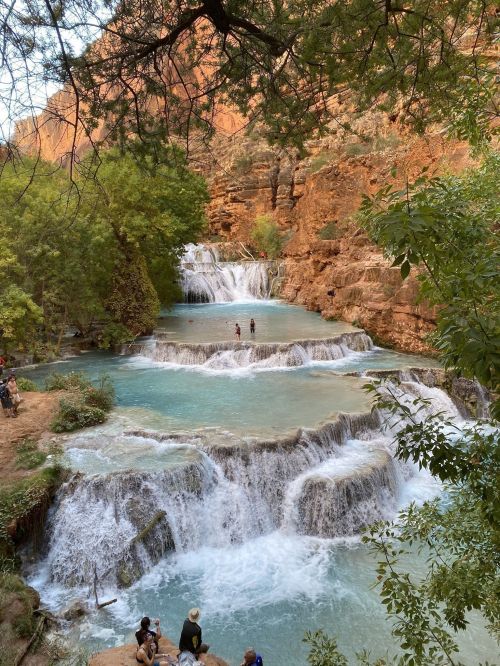 A scenic waterfall with turquoise pools, surrounded by lush greenery and red rock formations, with people enjoying the view.