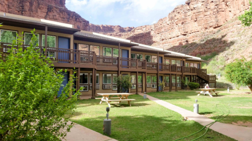 A two-story building with balconies surrounded by greenery and rocky cliffs, featuring picnic tables on a grassy area.